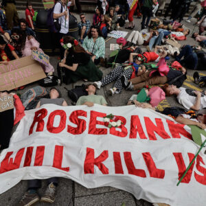 large white banner with words Rosebank will kill us in red on ground surrounded by people mostly lying round it