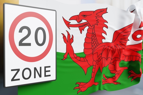 Road sign of 20mph zone and Welsh national flag in front of a street view. 3D illustration of the concept that Wales has lowered the speed limit on one-way roads in towns and cities