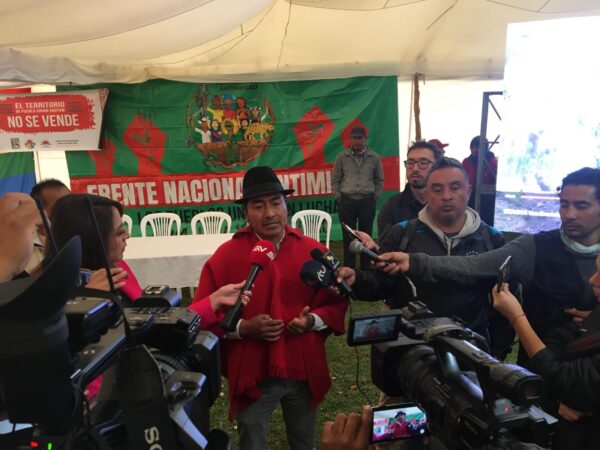 Leonidas Iza of CONAIE in front of FNA banner in red and green. Iza is in red poncho and black hat surrounded by lots of men in black with microphones