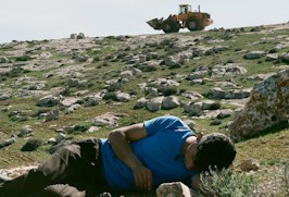 A brown boy lying in a green field full of rocks with a yellow tractor in the background.