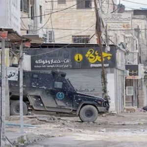 armoured car driving in front of closely packed buidlings one with sign in Arabic