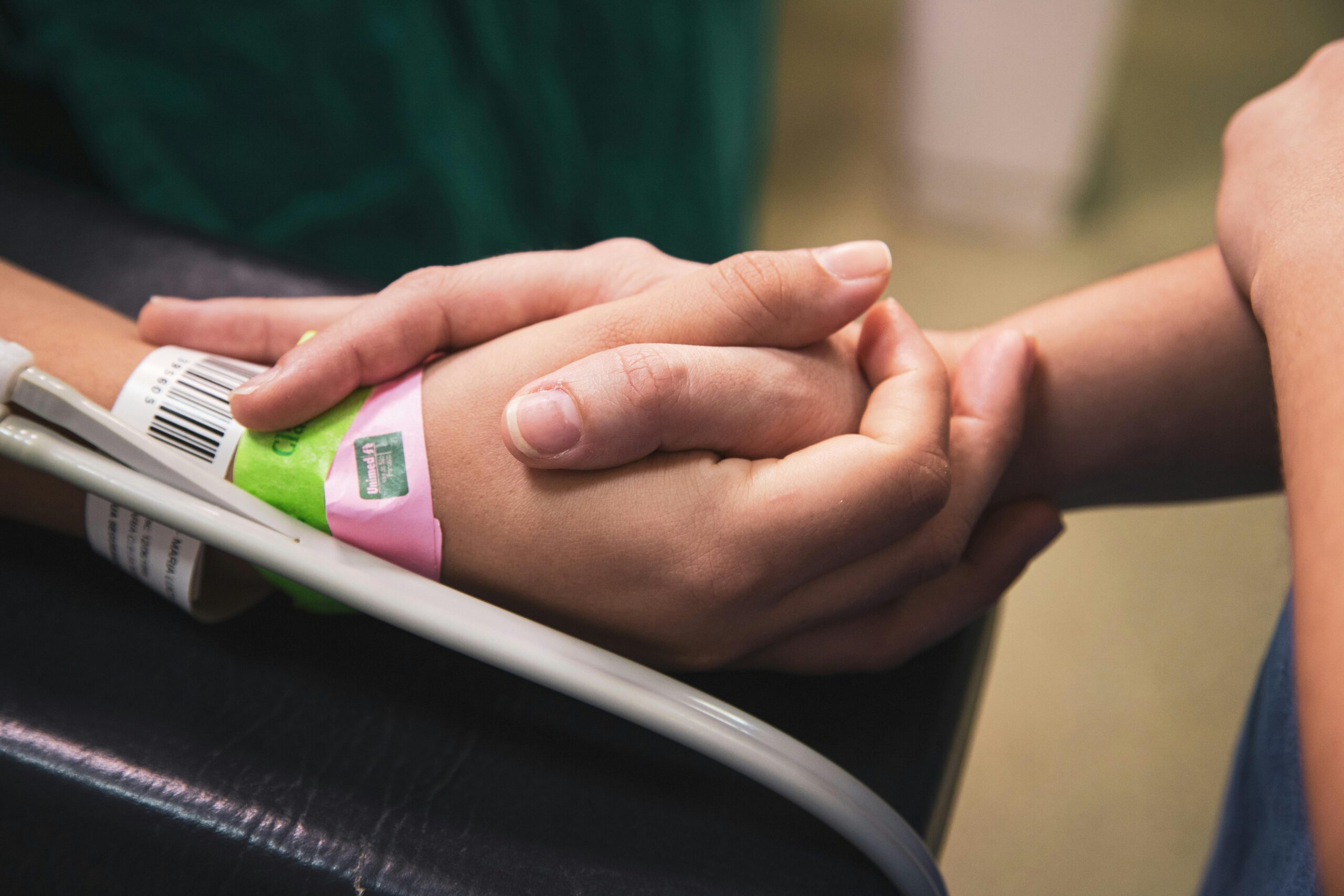 A close-up of two hands holding each other; one of the hands has wrist tags indicating that the person is likely a patient in a hospital. 
Photo by Hannah Barata