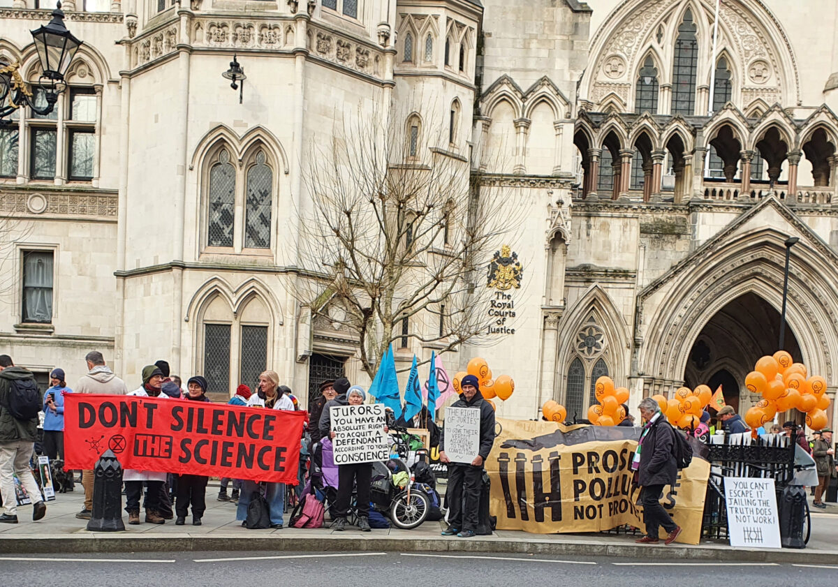 Landscaoe outside Royal courts of justuce with line of protestors and banners , Dont silence the science Black on red, others less easy to read, orange ballons and xr flags