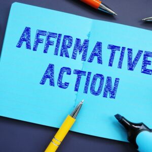 'Affirmative Action' written in dark blue on light blue pad lying on a desk and surrounded by various writing implements