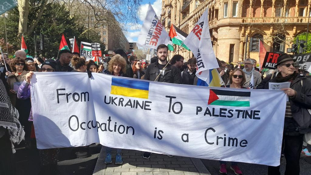 White Banner: From Ukraine to Palestine Occupation is a Crime with the respecyyive flags painted above the country names being carried by 5 or 6 people at front of photo with other people and banners further back