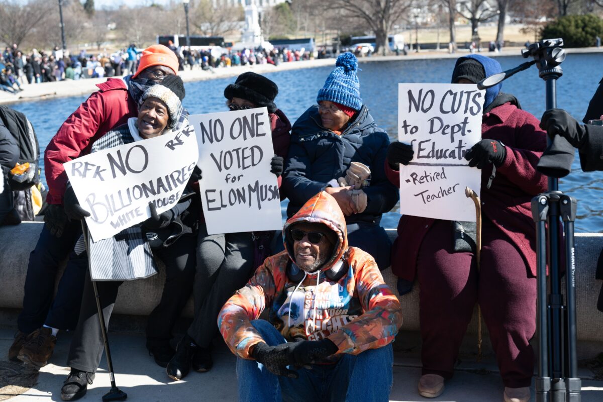 Group of African Americans in warm clothing with handmade signs, slogans include ' No one voted for Elon Musk' and 'No cuts to Dept of Education'.'