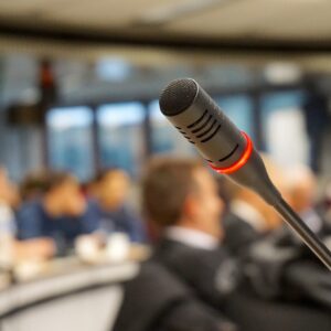A microphone at a meeting which is out of focus in the photo, with a red lit band around it to indicate that it is turned on.
