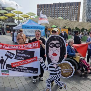 A group of activists hold up banners and placards at a disabled people's anti-cuts demo, with a cut out of Rachel Reeve's depicted as a thief.
