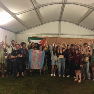A collection of young people inside a tent hold up a trans flag for a joyful group photograph