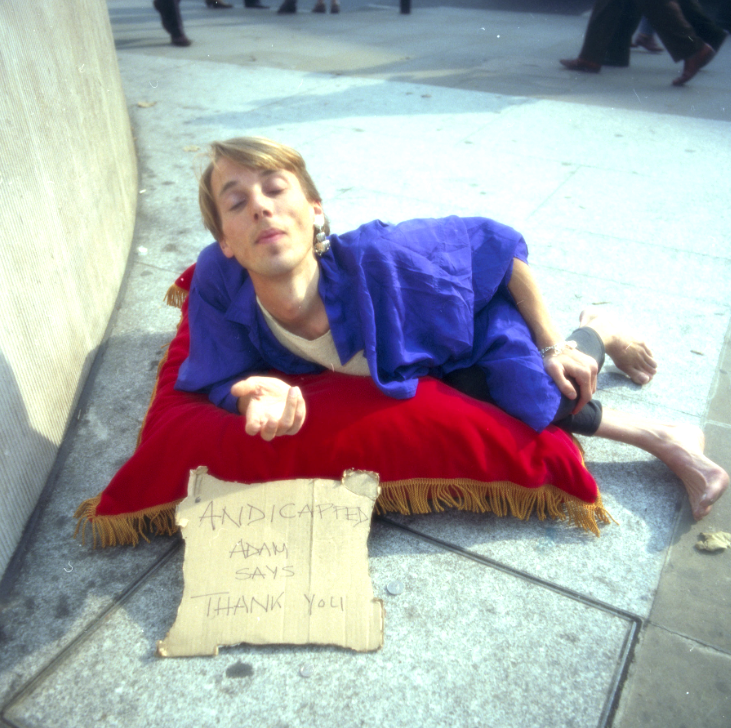 PErson lying on red cushion on ground with cardboard sign saying'Andicapped Adam says Thank You'