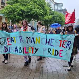 banner saying Geater Manchester Disability Pride in different coloured letters being carried by a number of people.