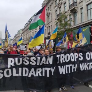 Black banner with test in whire - Russian troops out Solidarity with Ukraine with many Ukrainian and some Palestinian flags behind