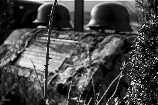 Two soldier's helmets sit on top of a grave marker with shrubs in the foreground, in a greyscale photograph.