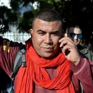 Photo of Lyes Touati looking at the camera wearing orange, talking into a mobile.