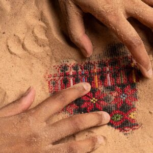 Two hands in sands uncovering intricate embroidery mianly in red and black typical of Palestinian work