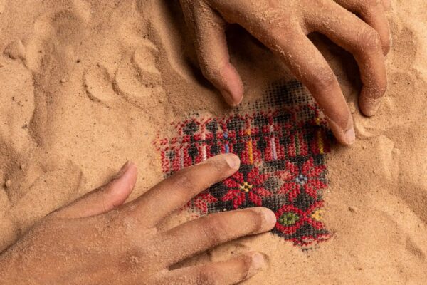 Two hands in sands uncovering intricate embroidery mianly in red and black typical of Palestinian work