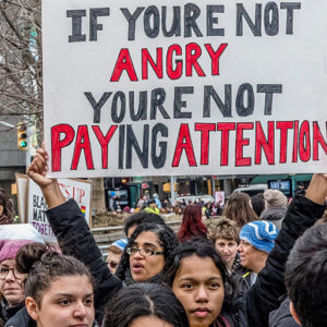 Large handmade banner 'If you are not angry you are not paying attention' being held aloft by young black woman surrounded by other women