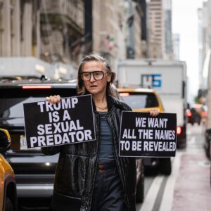 Person standing admist traffic with printed signs white out of black saying :Tump is a sexual predator and We want all the names to be revealed