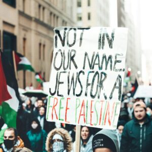 Masked protestor holds up a sign saying "Not in our name: Jews for a free Palestine" as oters march with palestinian flags.