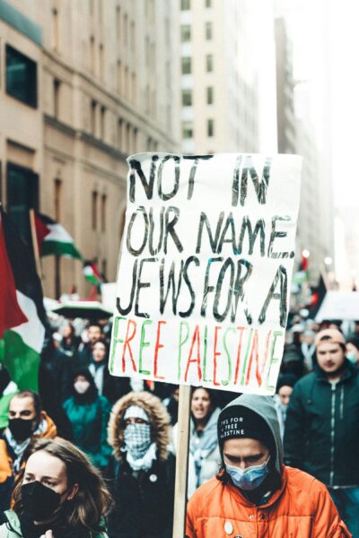 Masked protestor holds up a sign saying "Not in our name: Jews for a free Palestine" as oters march with palestinian flags.