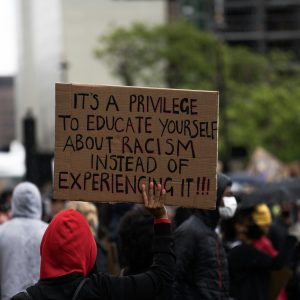 Protest where a person in a red hoodie holds up a board that reads "It's a privilege to educate yourself about racism instead of experiencing it!!!"