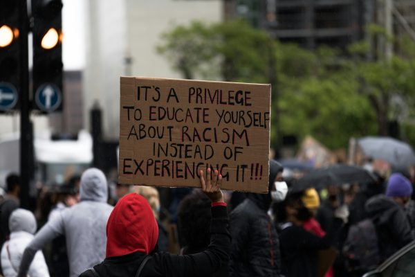 Protest where a person in a red hoodie holds up a board that reads "It's a privilege to educate yourself about racism instead of experiencing it!!!"