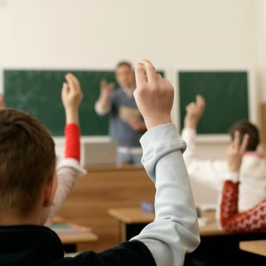 Students in a classroom raising hands, engaging in a lesson with the teacher.