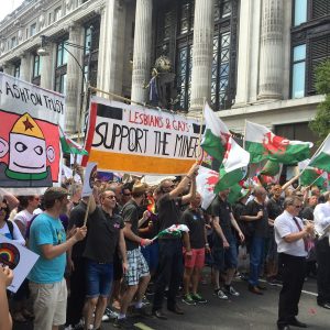 Group at Pride Parade holds up banners stating "Lesbians and Gays Support the Miners" and "Mark Ashton Trust"; some paraders hold up the flag of Wales.