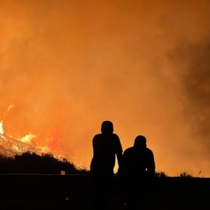 Silhouette of two people against a raging fire and an orange sky covered in smoke.